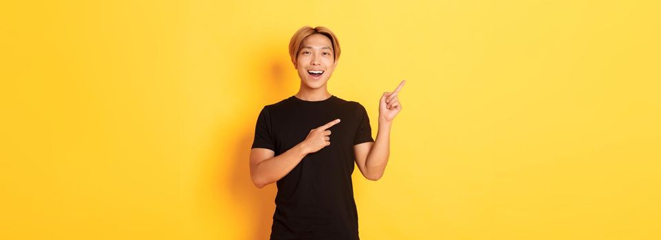 Portrait of cheerful asian man pointing fingers upper left corner, smiling and showing banner satisfied, yellow background.