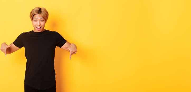 Portrait of excited and happy asian guy with blond haircut, pointing and looking down amused, standing yellow background.