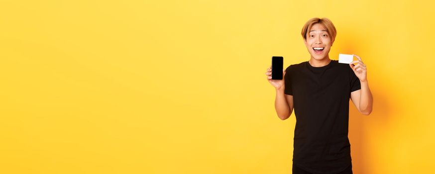 Portrait of excited happy asian man showing mobile phone screen and credit card with joyful smile, standing yellow background.