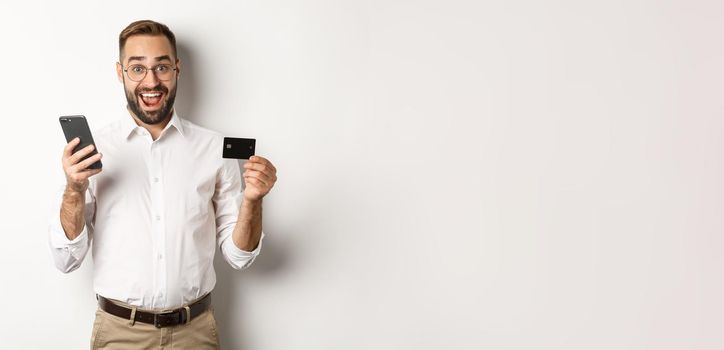 Business and online payment. Excited man paying with mobile phone and credit card, smiling amazed, standing over white background.