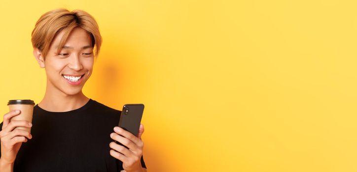 Close-up of handsome stylish asian male student, using mobile phone and drinking coffee. Guy messaging on smartphone, standing over yellow background.