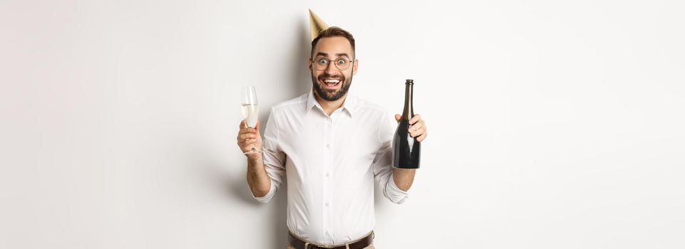 Celebration and holidays. Excited man enjoying birthday party, wearing b-day hat and drinking champagne, standing over white background.