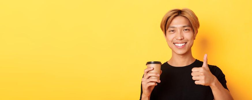 Close-up of satisfied asian handsome guy recommend cafe, holding cup of coffee and showing thumbs-up in approval, smiling pleased over yellow background.