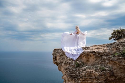 Blonde with long hair on a sunny seashore in a white flowing dress, rear view, silk fabric waving in the wind. Against the backdrop of the blue sky and mountains on the seashore