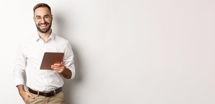 Confident business man holding digital tablet and smiling, standing against white background.