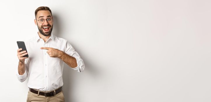 Man looking excited and pointing finger at mobile phone, showing good online offer, standing over white background.