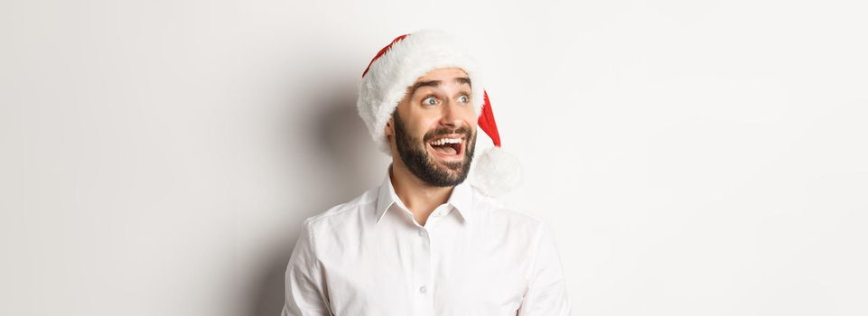 Close-up of amazed bearded man in santa hat, looking left with excitement, christmas promo offer, standing over white background.
