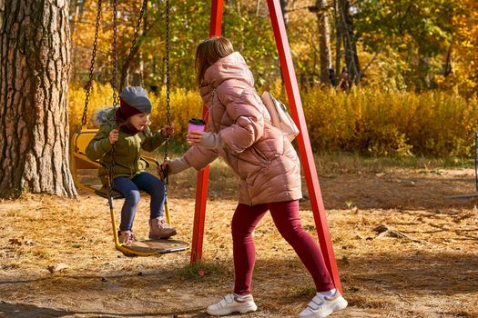 a woman in relation to her child or children. Young cute mother shakes her baby daughter on a swing in a bright autumn park.