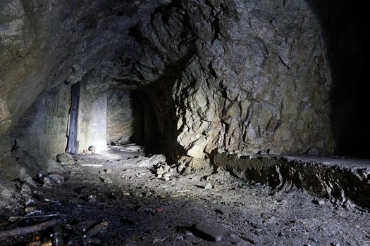 A dark artificial stone cave in the mountains. The light from the lantern falls on the rocks, where there are passages in the form of doors. Everything is wet. There are old branches and bricks.