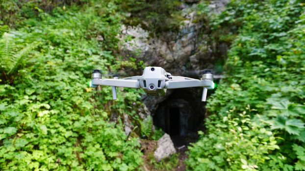 A quadcopter on the background of a green rocky gorge with the entrance to the cave. A gray drone hovers in air, red-orange lights glow, propellers spin. Blurred background with a view of tunnel path