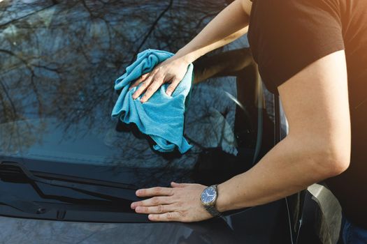 A man washes and wipes the windshield and body of his car with blue cloth rag.