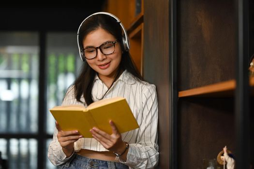 Portrait of college woman wearing headphone and reading book in library. People, knowledge and education concept.