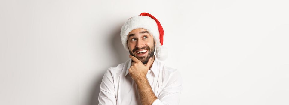 Party, winter holidays and celebration concept. Close-up of happy man planning christmas gift list, wearing santa hat, looking at upper left corner thoughtful, white background.