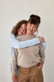 Young woman hugs happy mature smiling lady wearing casual clothing stand together in apartment room with beige wall closeup
