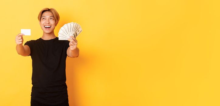 Happy good-looking asian guy looking thoughtful and pleased upper left corner while showing money and credit card, yellow background.