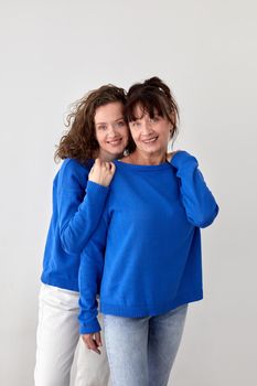 Glad loving mature mother and adult daughter wearing similar blue sweaters smile on gray background in studio