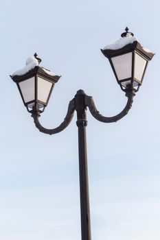 Vintage black street lamppost with two lamps covered with snow on blue sky background.