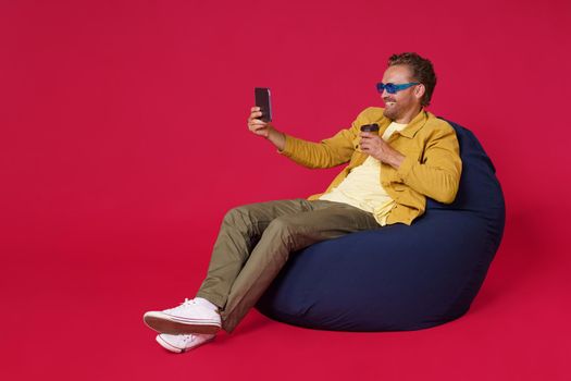 Handsome man 30s make selfie or video call using digital tablet drinking coffee from paper cup wearing casual denim outfit sitting on bag chair isolated on red background. Coffee shop concept.