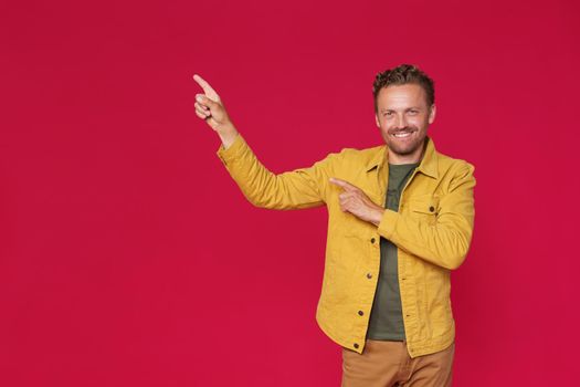 Happy bearded man in yellow jeans jacket pointing sideways up introducing copy space, mock up advertisement isolated on red background. Charming smiling freelancer introduce your business offer.