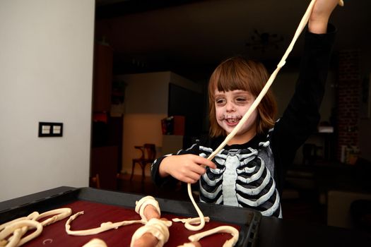 Cheerful kid in skeleton costume and makeup preparing pastries for baking on a baking sheet for the holiday halloween at home