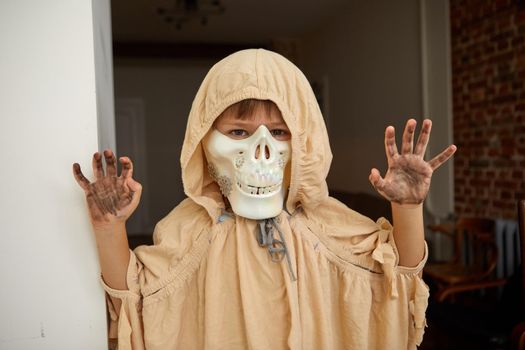 Child in spooky Halloween cape and skull jaw mask showing frightening gesture while looking at camera at home and having fun during holiday celebration