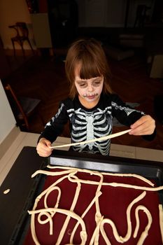 Boy child in costume and skeleton make-up preparing pastries for baking on a baking sheet for the holiday halloween at home