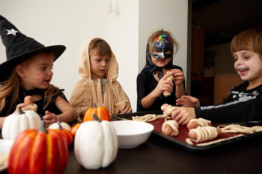 Company of content friendly kids in Halloween costumes gathering at table and prepare cooking sausage rolls during holiday celebration at home