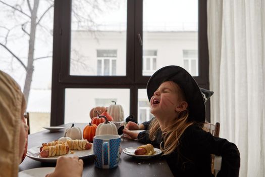 Cheerful child in costume of witch sitting at table and eating sausage roll while celebrating Halloween holiday at home and having fun