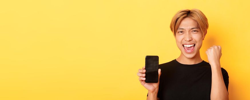 Close-up of happy rejoicing asian guy showing smartphone screen and saying yes, fist pump as triumphing, winning or achieve goal, yellow background.