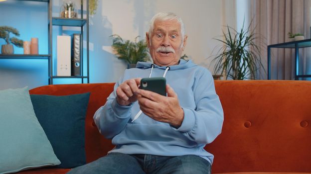 Happy excited joyful senior old grandfather man use mobile smartphone typing browsing say Wow yes great big win good news celebrate lottery jackpot. Elderly retired grandpa pensioner sit at home couch