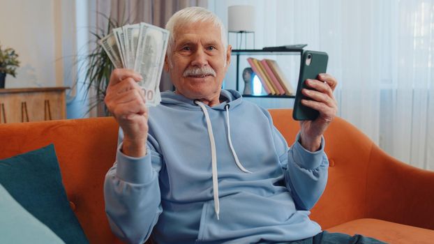 Planning family budget. Smiling senior old grandfather counting money cash, use smartphone calculate domestic bills at home. Joyful man satisfied of income and saves money for planned vacation, gifts