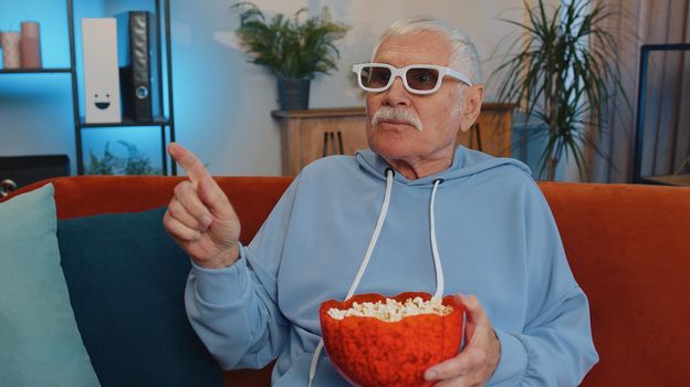 Excited elderly man on sofa eating popcorn and watching interesting tv serial, sport game, film, online social media movie content online at home. Senior grandfather enjoying domestic entertainment