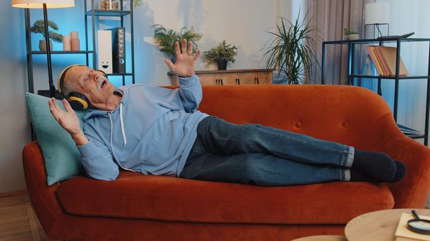 Senior old grandfather in wireless headphones relaxing lying on sofa at home choosing listening favorite energetic disco rock n roll music and dancing. People weekend activities. Happy overjoyed man