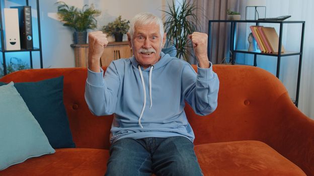 Happy old man shouting, celebrating success, winning, goal achievement, good news, raising fists in gesture I did it, lottery luck at home apartment. Senior grandfather indoors in living room on couch
