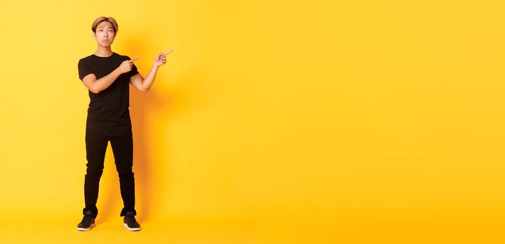 Full-length of sad and miserable asian guy in black clothes, pouting, pointing fingers upper right corner, standing yellow background.