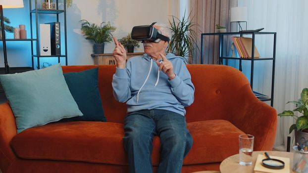 Senior old grandfather using virtual reality futuristic technology VR app headset helmet to play simulation 3D video game, watching film movie at modern home apartment. Man in goggles sitting on sofa