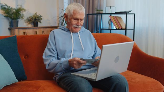 Planning budget. Rich happy senior grandfather counting money cash use laptop calculate domestic bills at home. Joyful elderly man satisfied of income earnings, saves money for planned vacation gifts