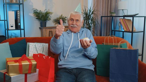 Happy senior grandfather wears festive birthday hat hold cupcake makes wish joyful congratulating blowing burning candle on cake. Mature man celebrating anniversary party on couch with gifts at home