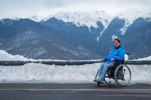 A happy woman dressed in a blue coat sits in a wheelchair on a point view and looking at the snow-capped mountains