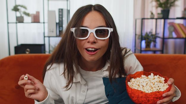 Excited adult girl sitting on sofa eating popcorn and watching interesting tv serial, sport game, film, online social media movie content online at home. Young woman enjoying domestic entertainment