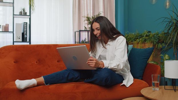 Overjoyed caucasian girl at home sitting on couch with laptop scream in delight raise hands in triumph winner gesture celebrate success win money in lottery. Excited young woman get online good news