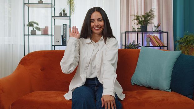 Portrait of caucasian adult girl smiling friendly at camera and waving hands gesturing hello, hi, greeting or goodbye, welcoming with hospitable expression. Young woman indoor at home in room on couch