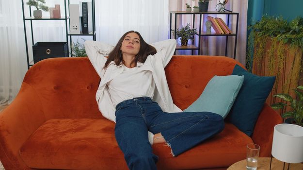 Tired caucasian adult girl lying down on couch taking a rest at home. Carefree young woman napping, falling asleep on comfortable orange sofa with pillows. Closed her eyes enjoy daytime nap alone