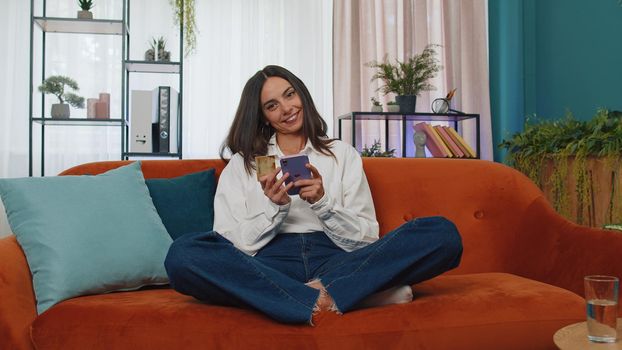 Happy shopaholic consumer young woman sitting at modern home apartment indoors making online order food delivery, payment with credit plastic bank card. Discount sale on internet. Adult girl on couch