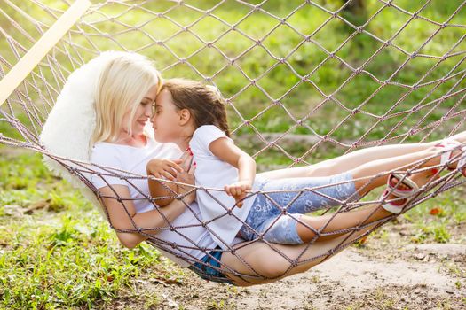 Happy mother and daughter relaxing together in a hammock at garden in sunny summer day. Family playing in hammock.