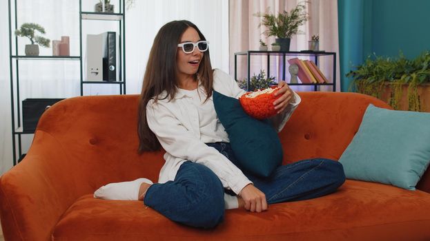 Excited adult girl sitting on sofa eating popcorn and watching interesting tv serial, sport game, film, online social media movie content online at home. Young woman enjoying domestic entertainment