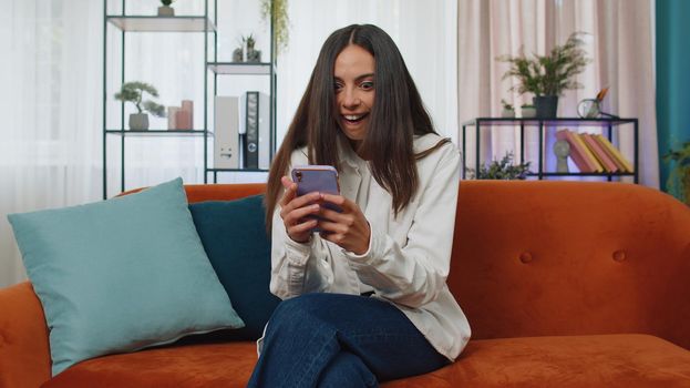 Portrait of lovely winner girl use mobile smartphone typing browsing say Wow yes found out great big win good news celebrate, lottery jackpot. Young woman at home in living room sitting on orange sofa