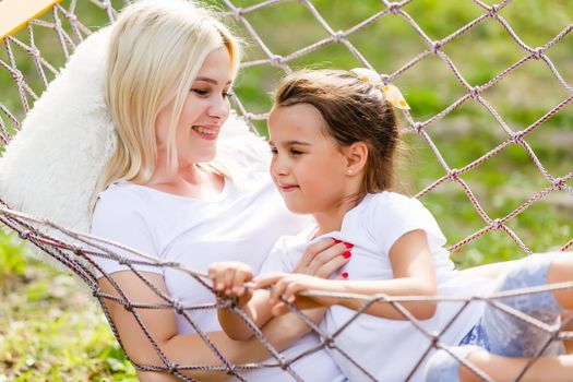 Happy mother and daughter relaxing together in a hammock at garden in sunny summer day. Family playing in hammock.