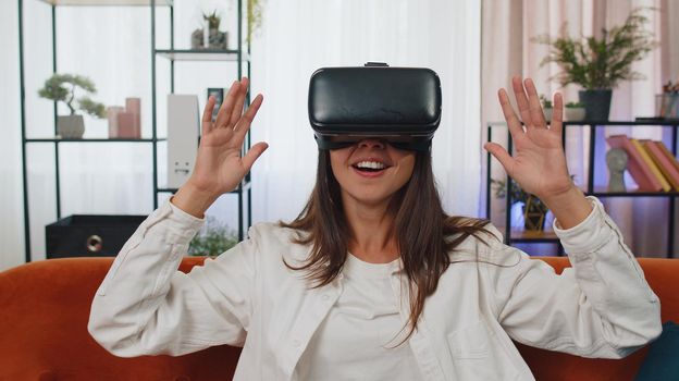 Young Caucasian girl wearing VR headset resting at home sitting on sofa in living room alone using futuristic technology making gestures with hands in air, play 3D video game. Virtual reality glasses