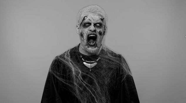 Sinister man in carnival costume of Halloween crazy zombie with bloody wounded scars face screaming, shouting, trying to scare on gray. Horror theme of cosplay wounded undead, beast, monster